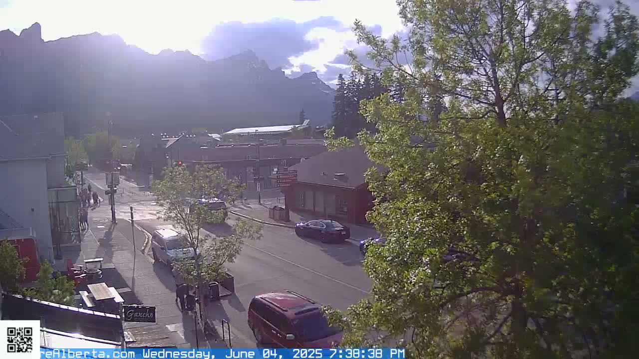A partly cloudy afternoon scene shows a street in a town nestled in a valley with mountains in the background.