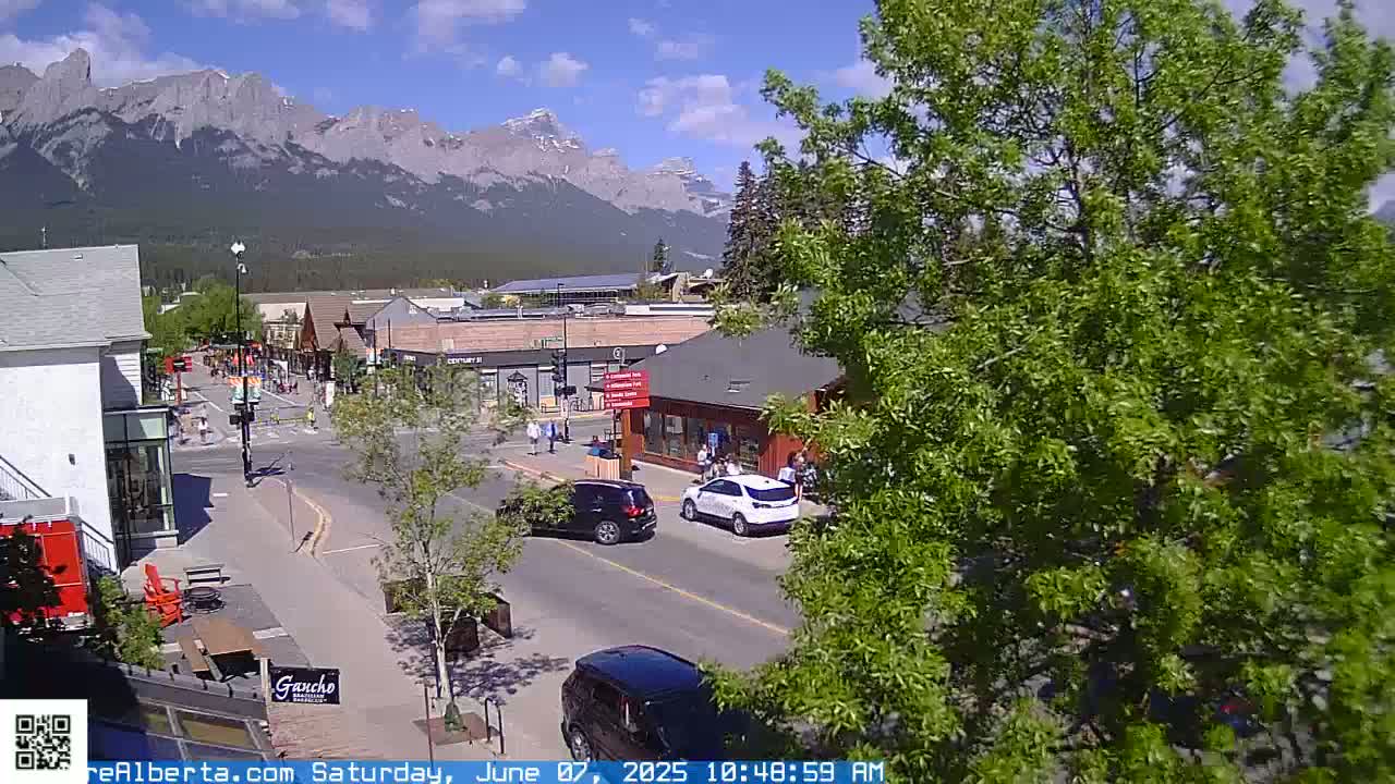 A sunny, mountain-view town square scene with cars parked and driving on the street, pedestrians walking, and lush green trees.
