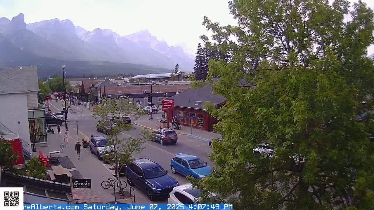 A partly cloudy afternoon scene shows a mountain range in the distance behind a town's street with cars, pedestrians, and businesses.