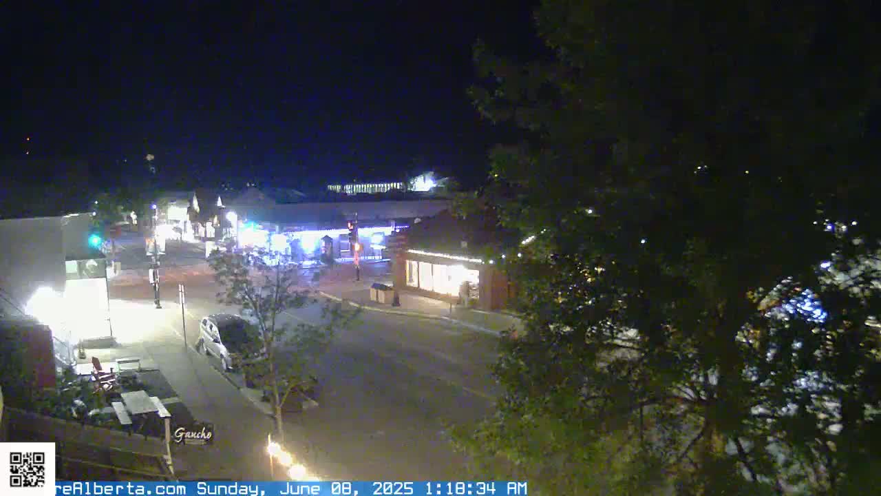 A nighttime, street-level view of a sparsely populated town square with several lit buildings and a few vehicles parked along the streets.