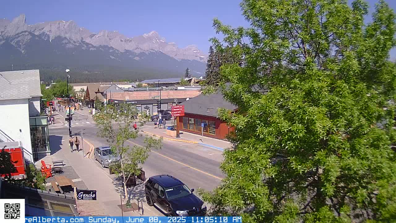 A sunny day in a mountain town shows a street scene with cars, pedestrians, and buildings, with a mountain range visible in the background.