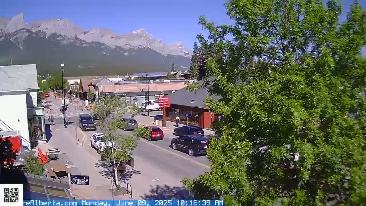 A sunny day in a mountain town shows a street with cars and pedestrians, with a mountain range in the background.