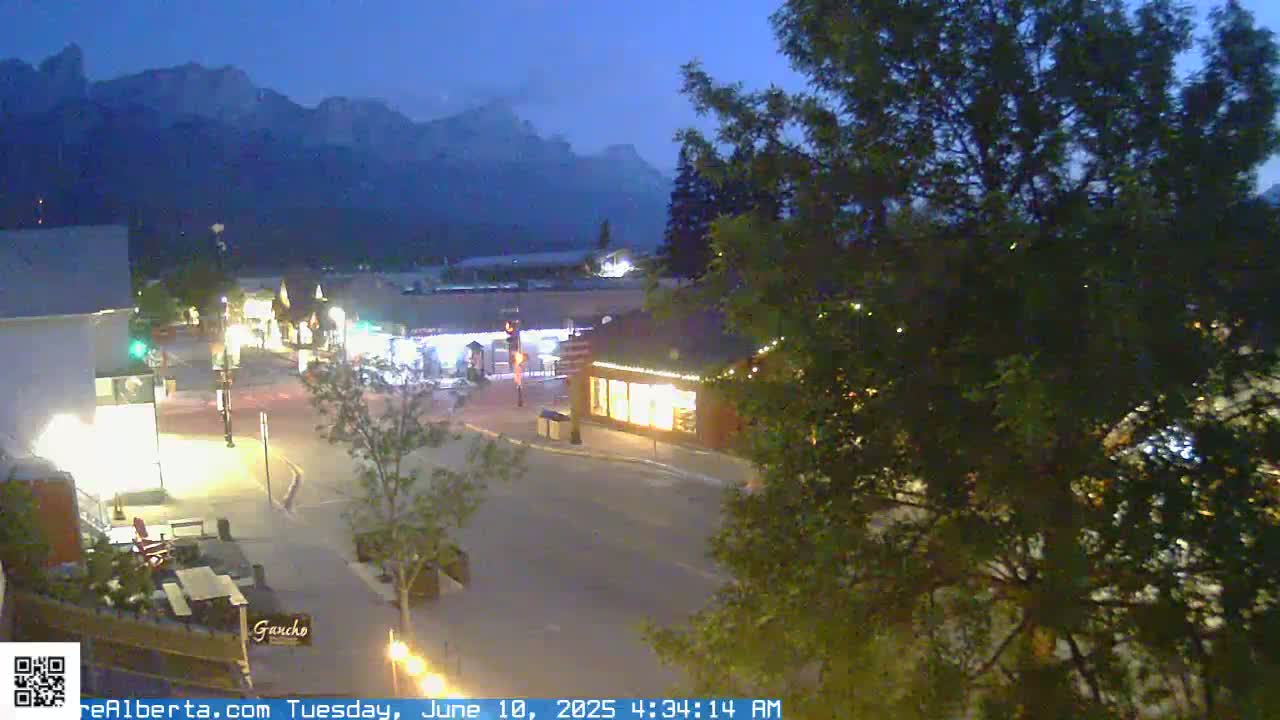 A nighttime view of a town square with dimly lit buildings and a mountain range in the background under a twilight sky.