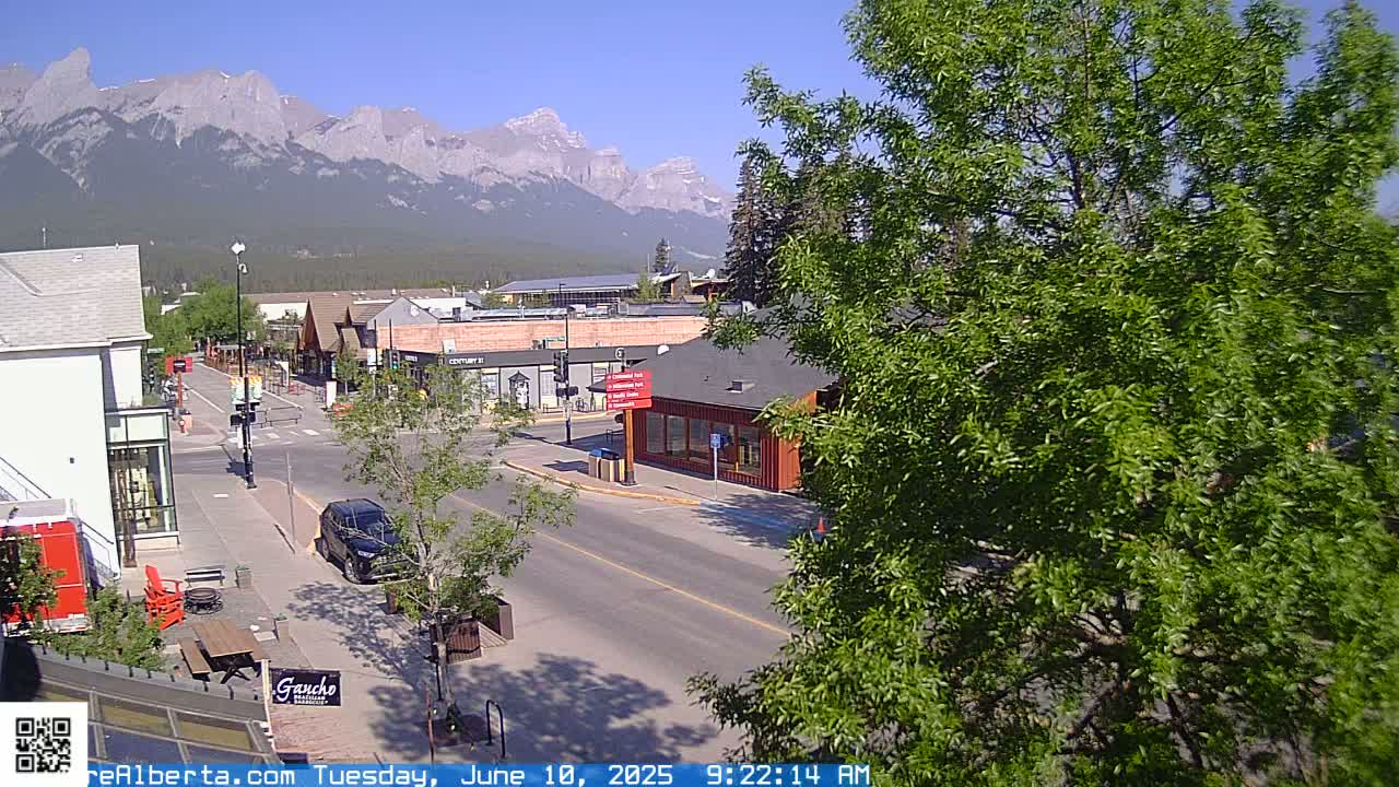 A street scene in a town nestled against a mountain range under a clear, sunny sky shows buildings, parked cars, and lush green trees.