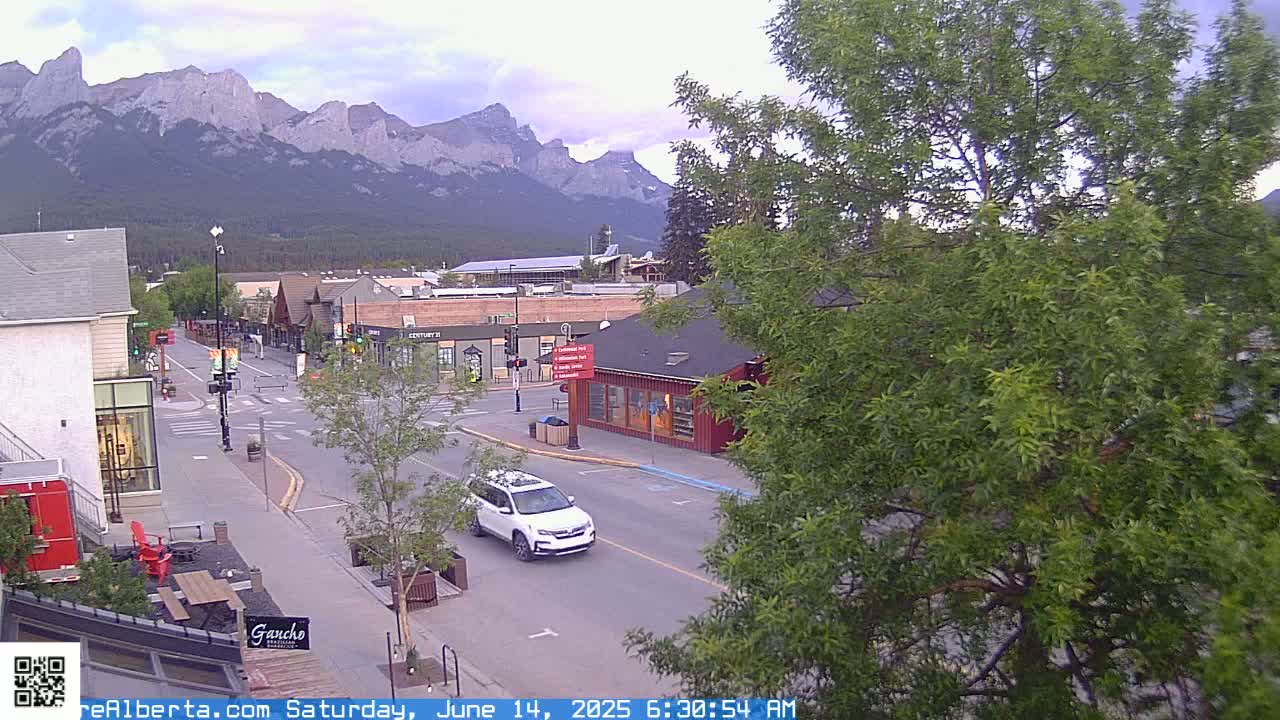 A partly cloudy morning view of a mountain town street scene, with a white SUV driving down the road, and mountains visible in the background.
