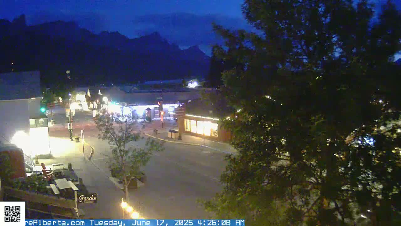 A nighttime view of a town square with low-level lighting, bordered by buildings and trees, under a dark sky with mountains in the background.