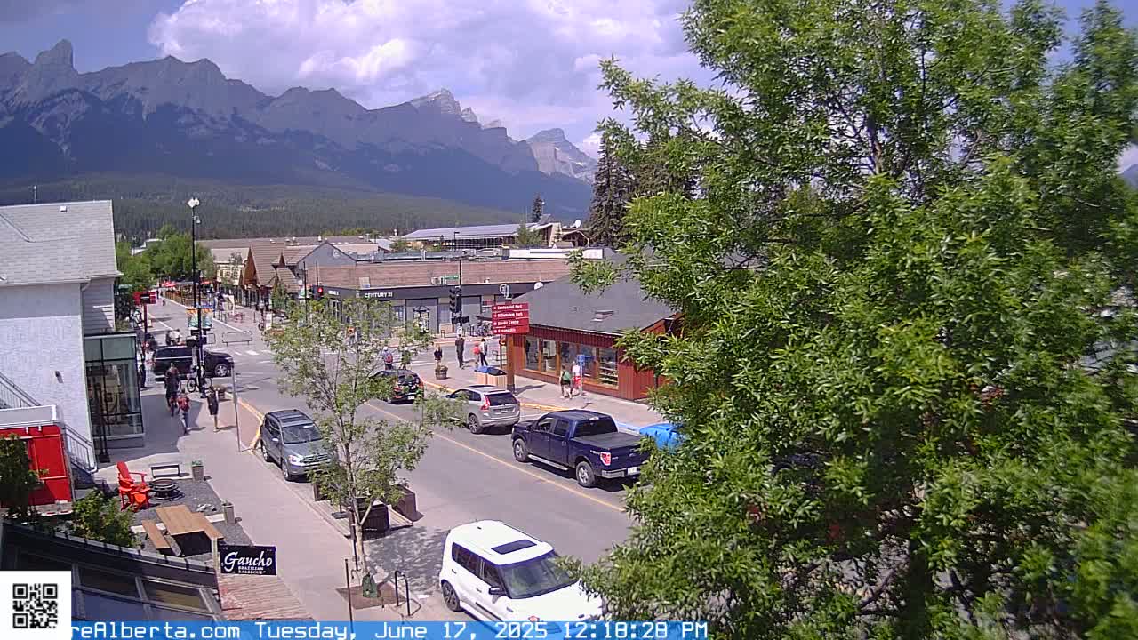 A partly cloudy day reveals a mountain range in the background of a street scene with cars parked along the sides of the road and several pedestrians walking.