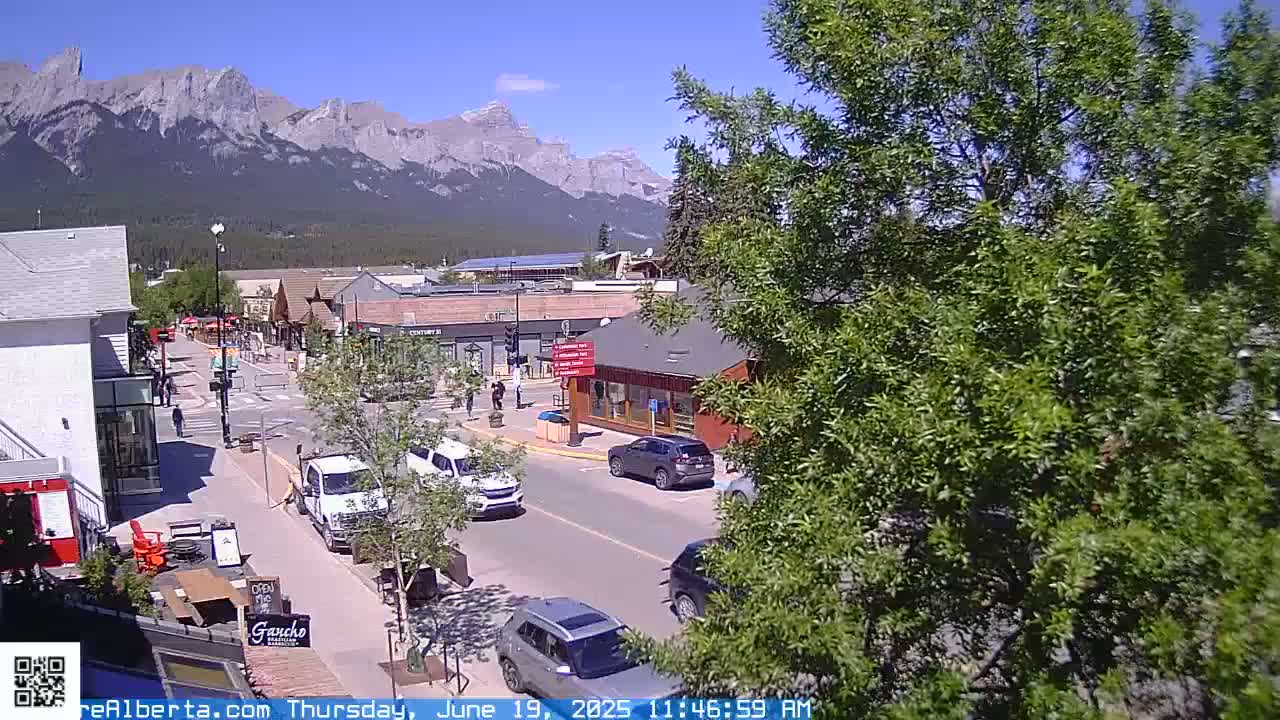 A sunny day view of a mountain town street scene with cars parked and driving, shops and buildings lining the road, and a mountain range in the background.