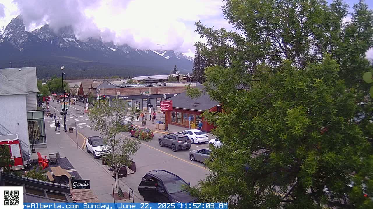 A partly cloudy day reveals a mountain range in the distance behind a street scene of cars and pedestrians in a town with various shops and buildings.