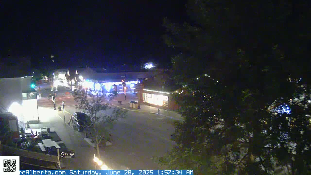 A nighttime, overhead view of a sparsely populated street in a town, with a few buildings illuminated and some trees visible in the foreground.