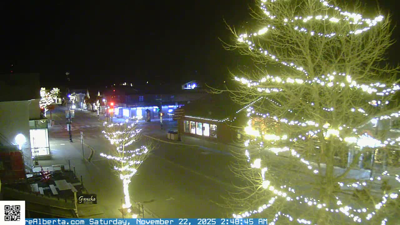 A quiet town street at night is brightly illuminated by decorative lights strung on bare trees and buildings, under clear weather conditions.
