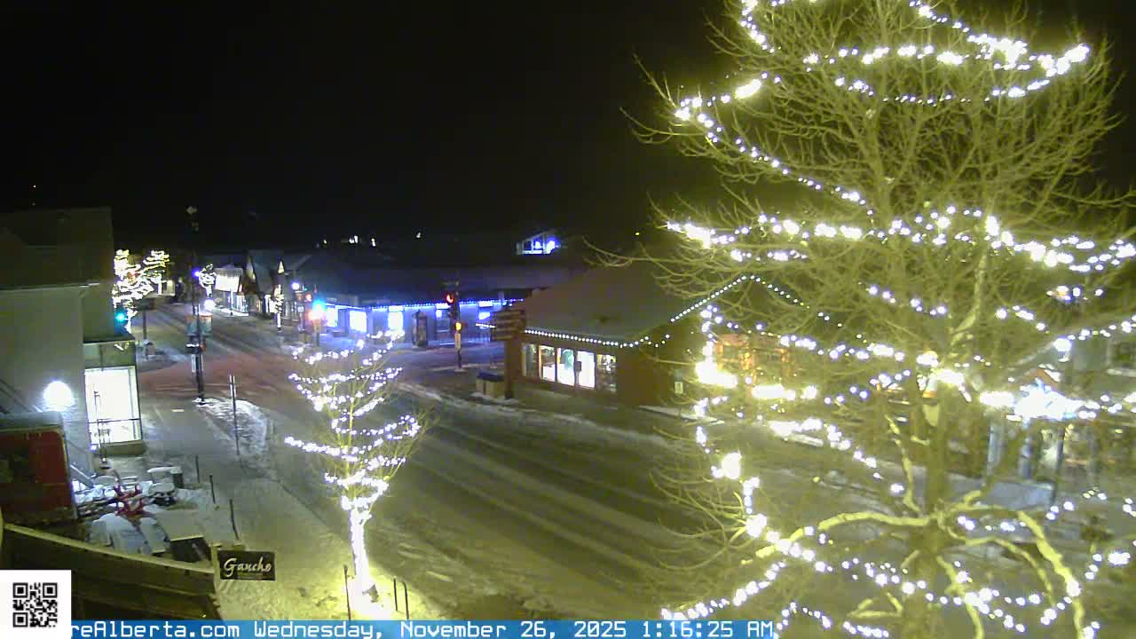 A snow-covered street in a festive town at night is brightly illuminated by white string lights on bare trees and buildings under clear, cold conditions.