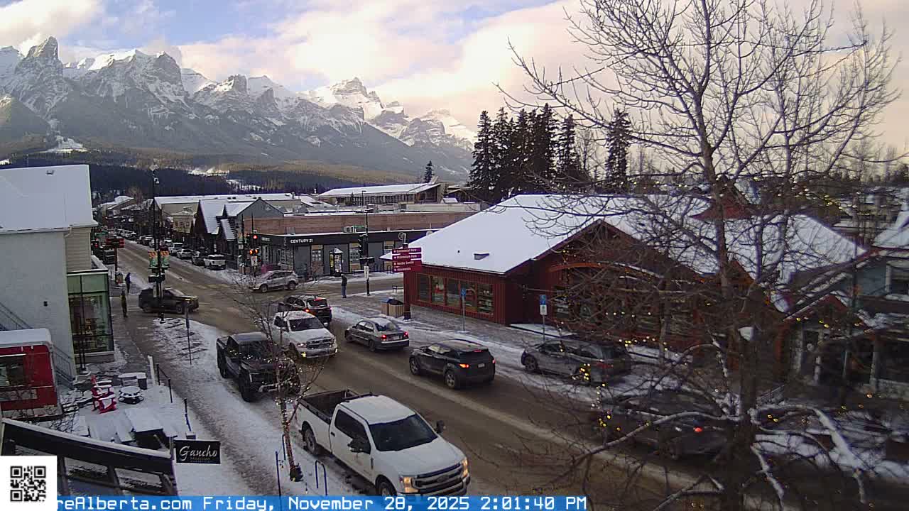 A wintery scene depicts a busy town street covered in snow, lined with buildings and numerous cars, set against a backdrop of majestic snow-capped mountains under a partly cloudy sky.