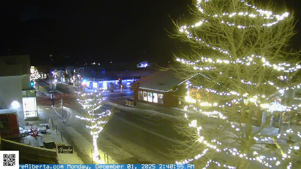 A snowy street scene at night is brightly illuminated by numerous white string lights adorning bare trees and the rooftops and facades of buildings, indicating clear, cold winter weather.