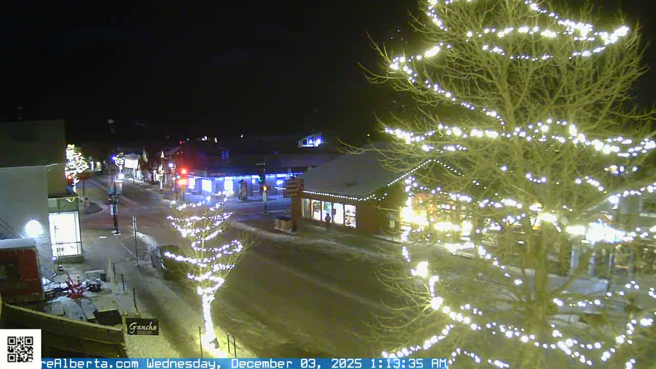 A clear and cold winter night illuminates a festive street lined with snow-covered buildings and bare trees brightly decorated with white lights.