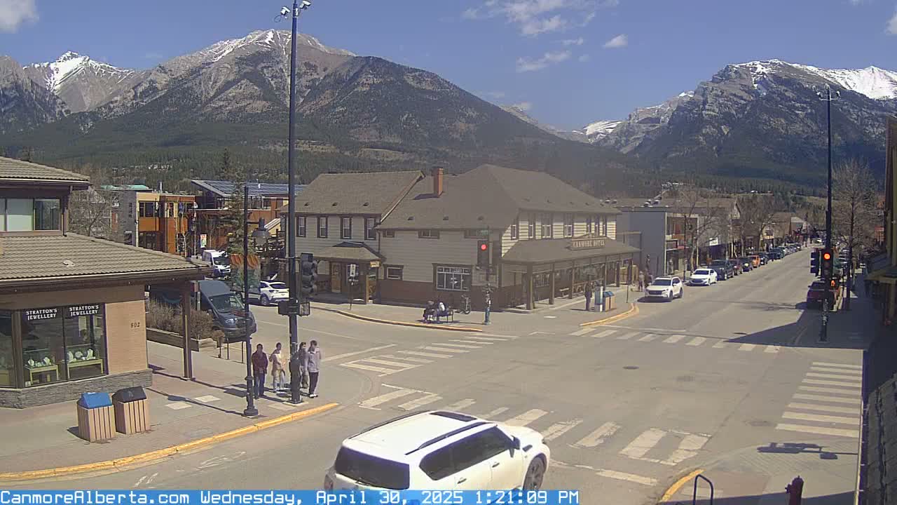 A sunny day in a mountain town shows a street intersection with several pedestrians and cars, with snow-capped mountains in the background.