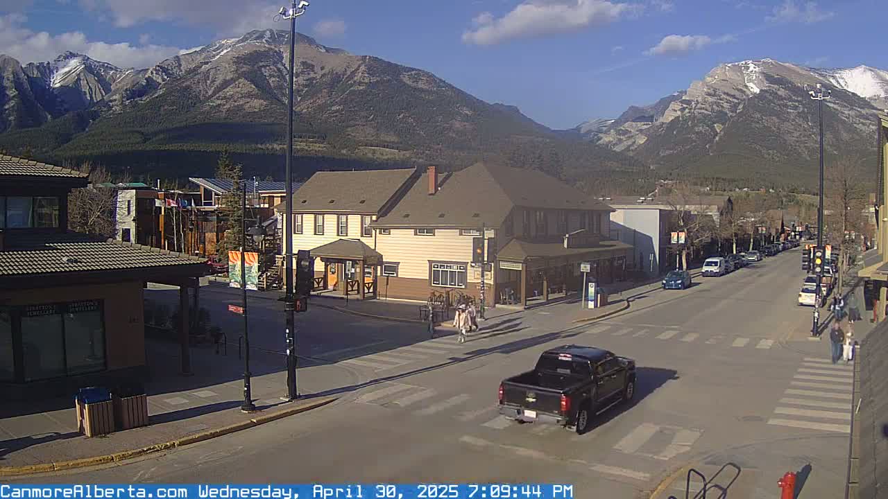 A sunny day reveals a mountain town street scene with buildings, vehicles, and pedestrians, all set against a backdrop of majestic mountains.