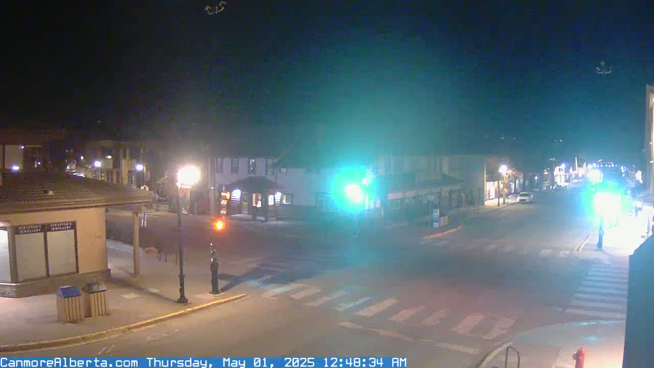 A nighttime street scene shows a mostly empty intersection in a small town with streetlights illuminating buildings and crosswalks.