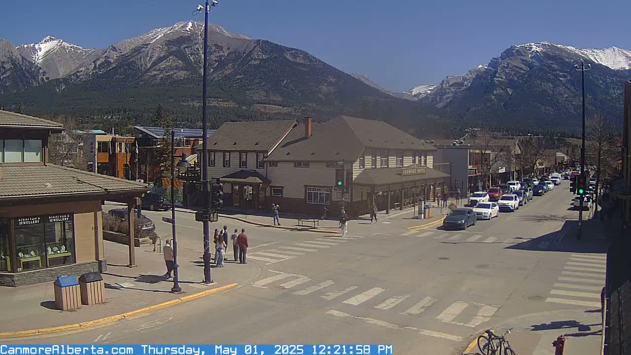 A sunny day reveals a mountain town scene with pedestrians crossing a street in front of shops and a hotel, with cars parked along the roadside and mountains in the background.