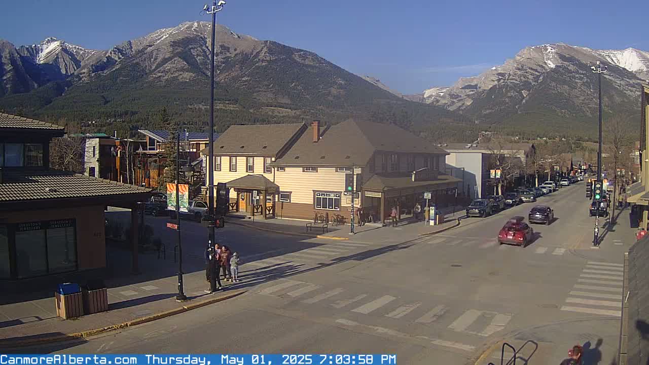 A sunny street scene shows a town nestled in a valley with mountains in the background, and several cars and pedestrians near a crosswalk.