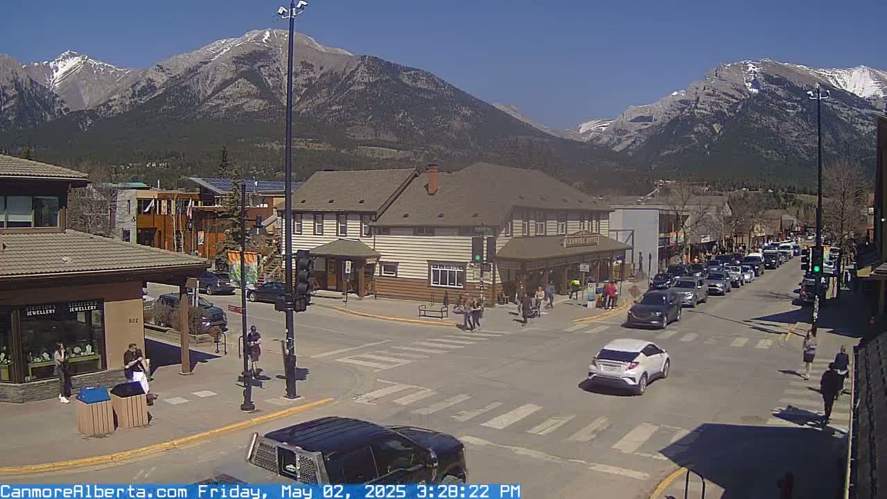 A sunny day in a mountain town shows a street intersection with cars and pedestrians, buildings lining the street, and snow-capped mountains in the background.
