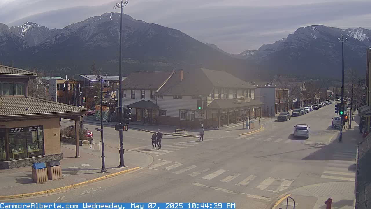 A street scene in a town nestled against a mountain range under a mostly cloudy sky shows pedestrians crossing a street, cars driving by, and several buildings lining the street.