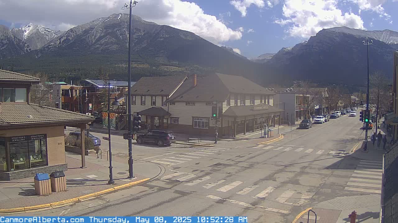 A sunny street scene in a mountain town shows several cars driving past shops and a hotel, with snow-capped mountains in the background.