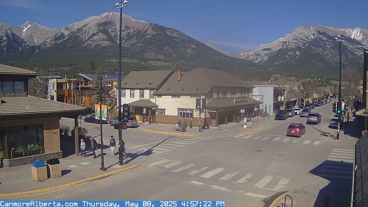 A sunny day reveals a town's street intersection with pedestrians and cars, nestled against a backdrop of majestic mountains.