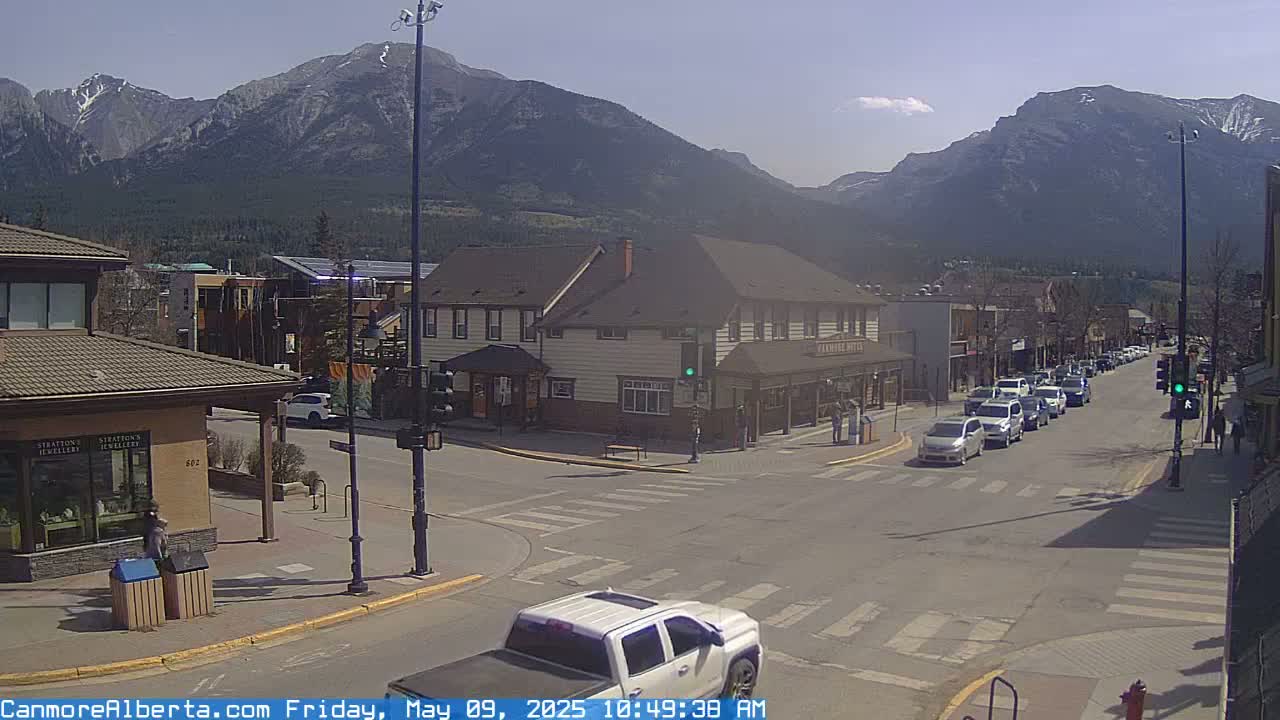 A sunny mountain town street scene shows several vehicles at a crosswalk in front of buildings, with a mountain range in the background.