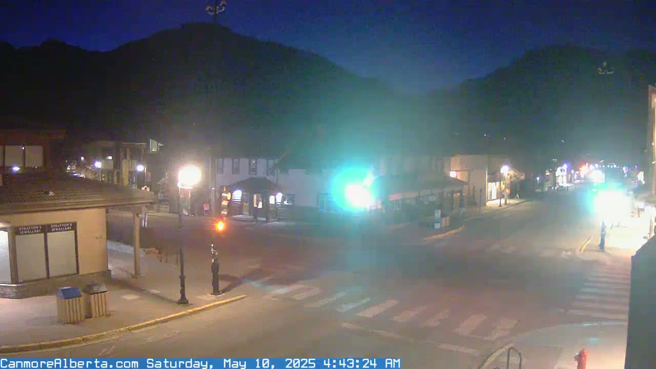 A nighttime street scene shows a mostly empty intersection in a town at the base of a dark mountain under a clear, dark sky.