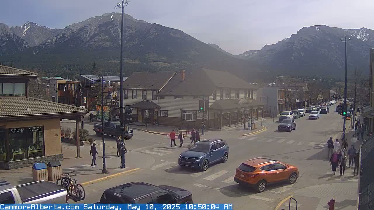 A sunny mountain town scene shows a street intersection with pedestrians and cars, in front of buildings and with mountains in the background.