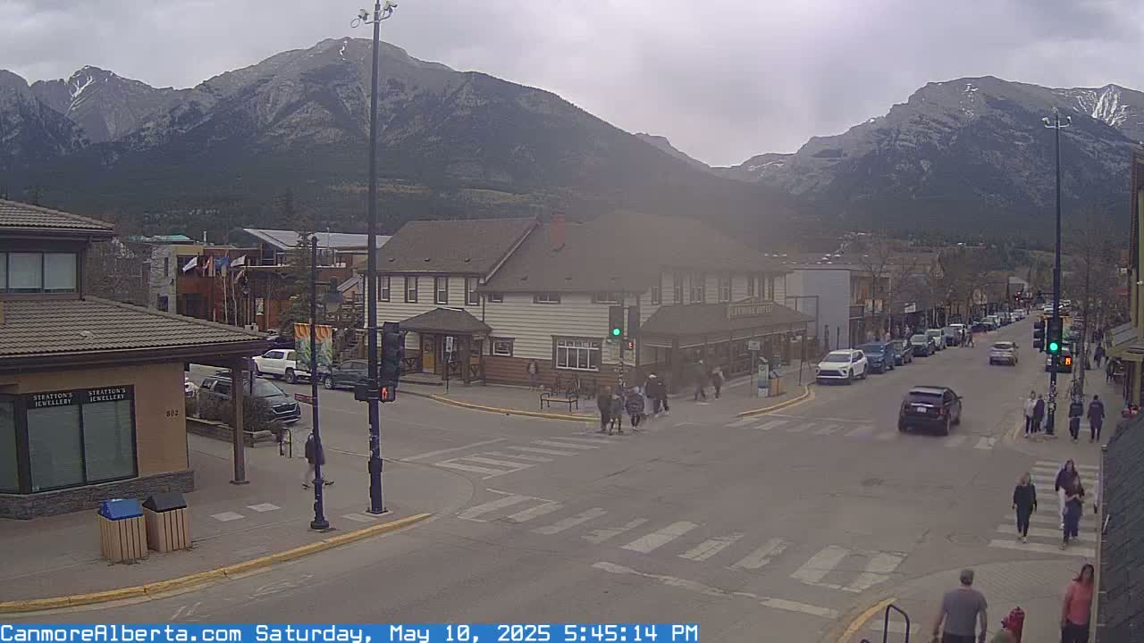 A street scene shows a town with several cars parked and driving on the street, pedestrians crossing at a crosswalk, and mountains in the background under an overcast sky.