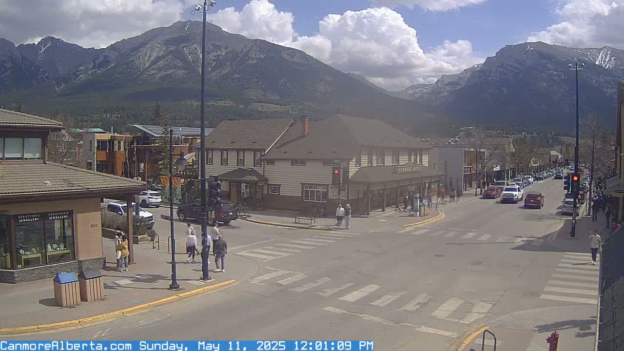 A partly cloudy day in a mountain town shows a street intersection with pedestrians and cars, with shops and buildings lining the street and mountains in the background.