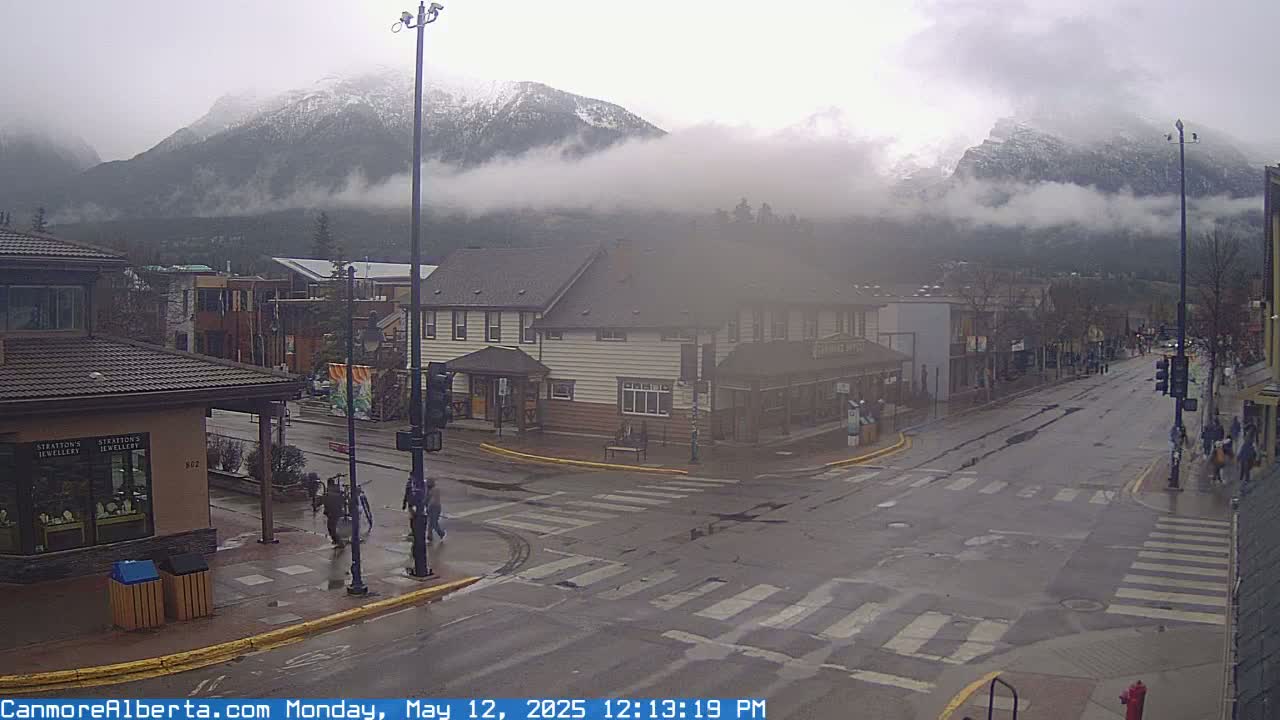A wet street scene shows a town intersection with pedestrians and buildings, with snow-capped mountains partially obscured by fog in the background.