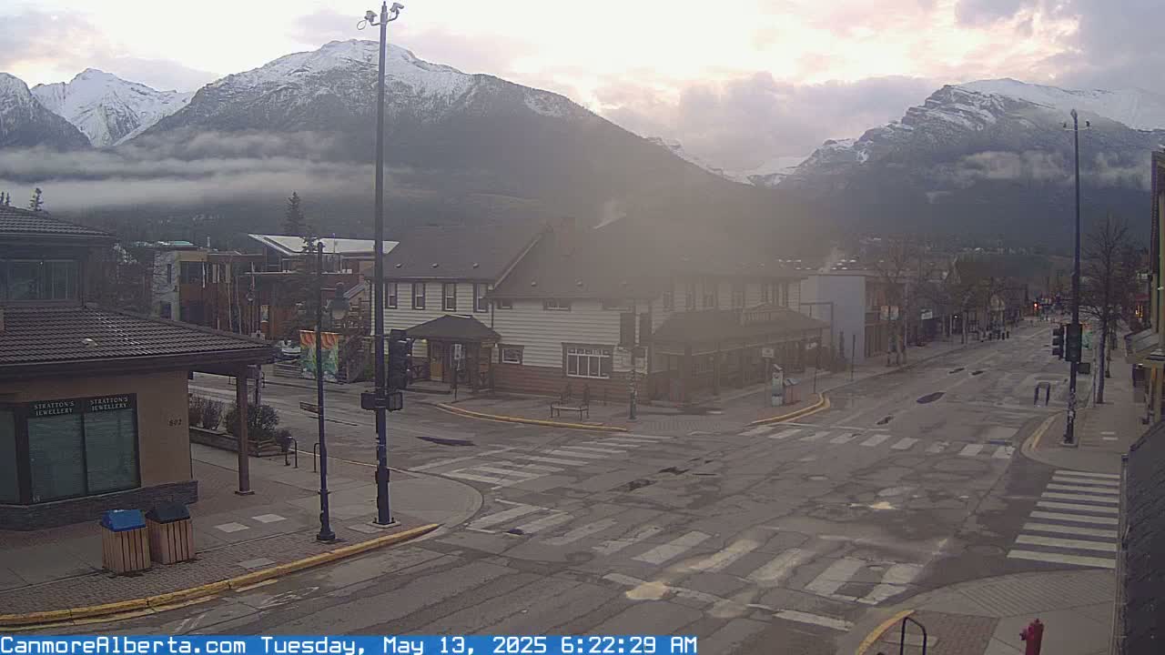A town's street intersection with snow-capped mountains in the background under a partly cloudy sky.