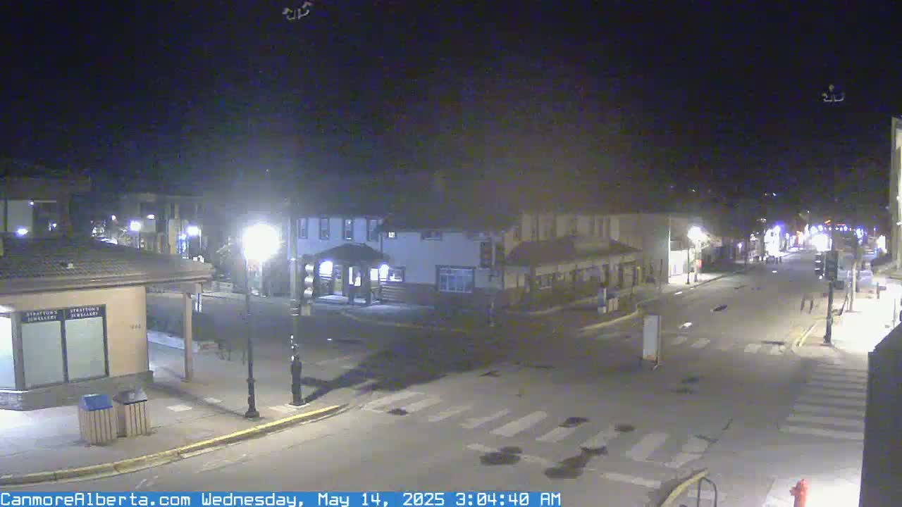 A nighttime, mostly empty street scene in a small town features several buildings under streetlights, with a few pedestrians visible.