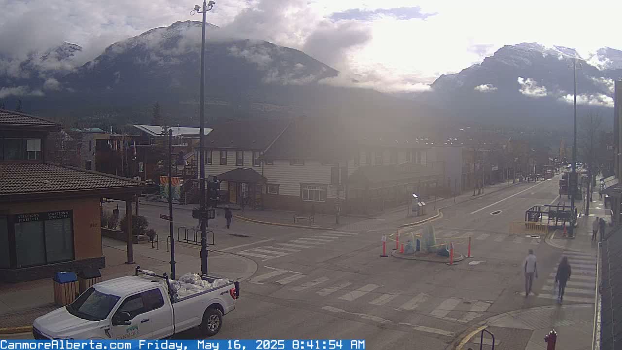 A partly cloudy day reveals snow-capped mountains in the distance, overlooking a town square with a few pedestrians and a white pickup truck.