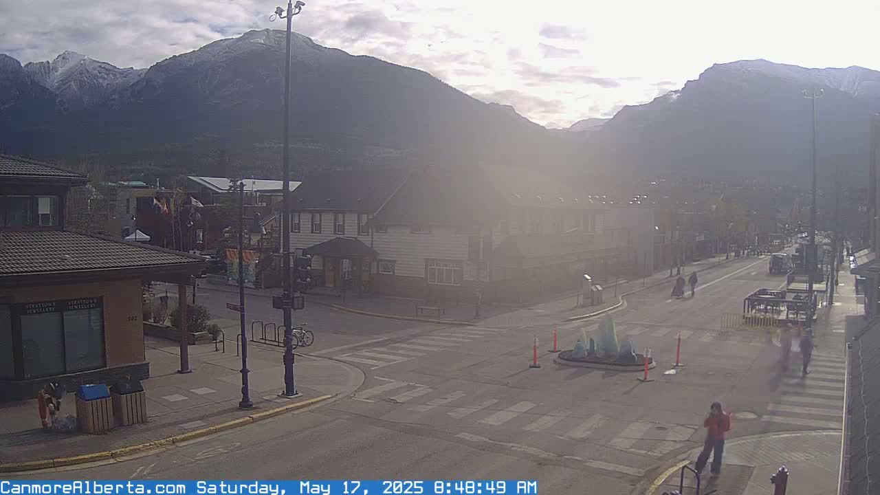 A hazy, sunny day reveals a mountain range behind a town's street intersection, where a few pedestrians and a bicycle are visible near buildings.