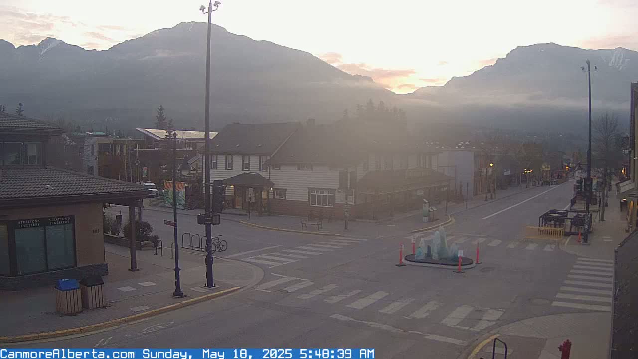A hazy sunrise illuminates a quiet town street nestled in a valley, with mountains shrouded in mist in the background.