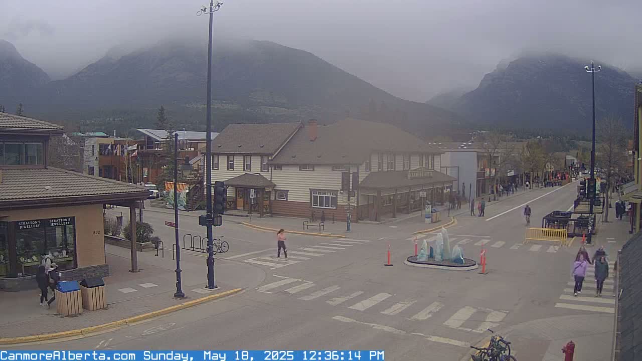 A mostly cloudy day reveals a mountain range behind a town's main street where people are walking and biking, a fountain sits in the middle of a crossroads.