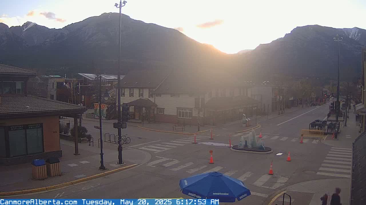 A hazy sunrise illuminates a mountain range behind a quiet town square with a few pedestrians and a partially closed-off intersection.