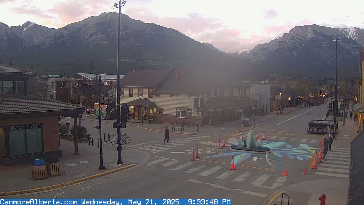 A town square with a fountain surrounded by construction barriers is situated at the foot of a mountain range under an overcast sky.