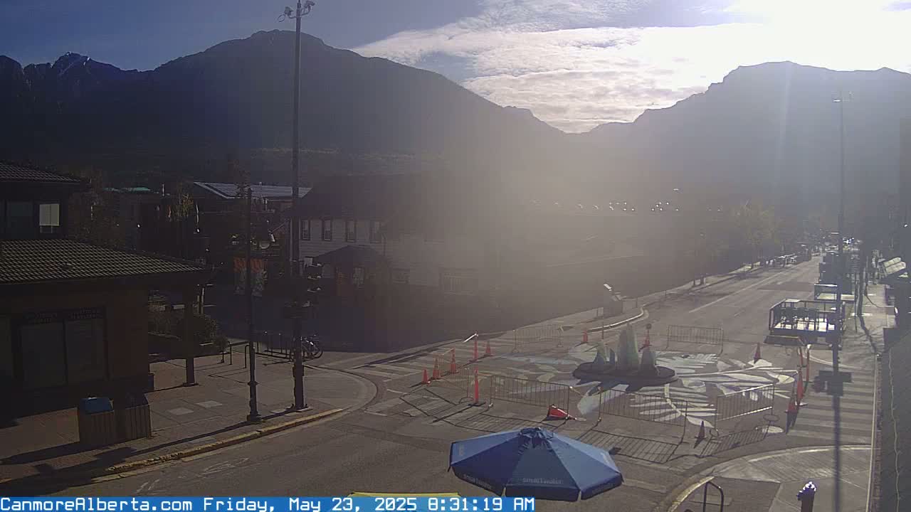 A hazy sunrise illuminates a mountain range behind a town square with a street and buildings, partially obstructed by barricades and a blue umbrella.