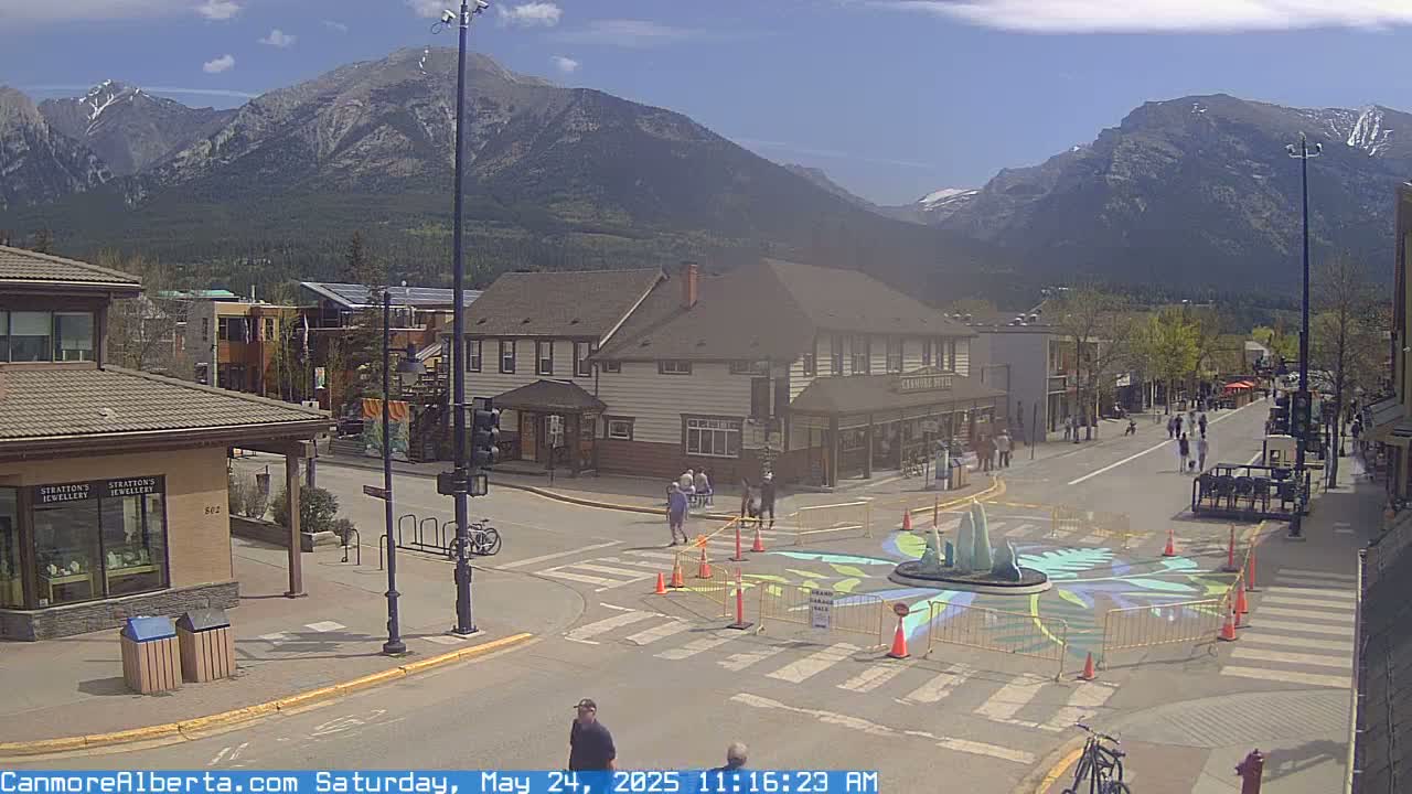 A sunny day in a mountain town shows a street scene with pedestrians and buildings, including a large hotel, with a central roundabout featuring a fountain and colorful street art.