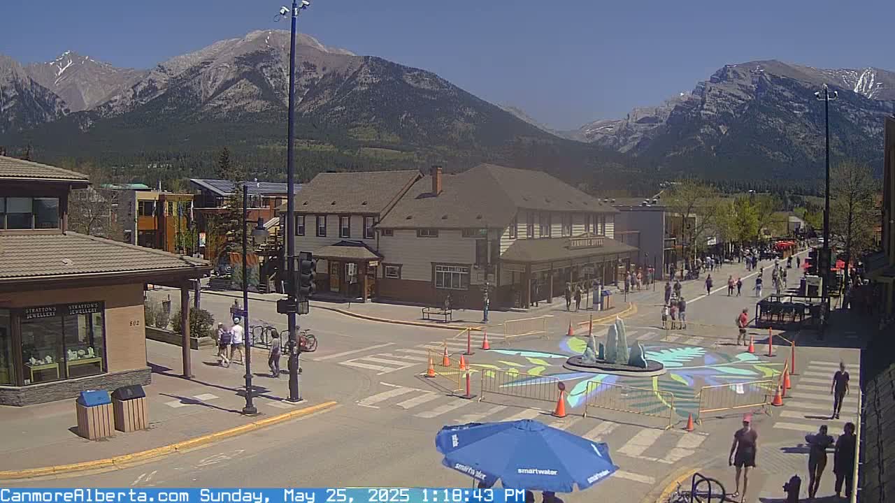 A sunny town square, surrounded by buildings and mountains, features a pedestrian crossing with a painted design and a fountain in the center, with people walking around.