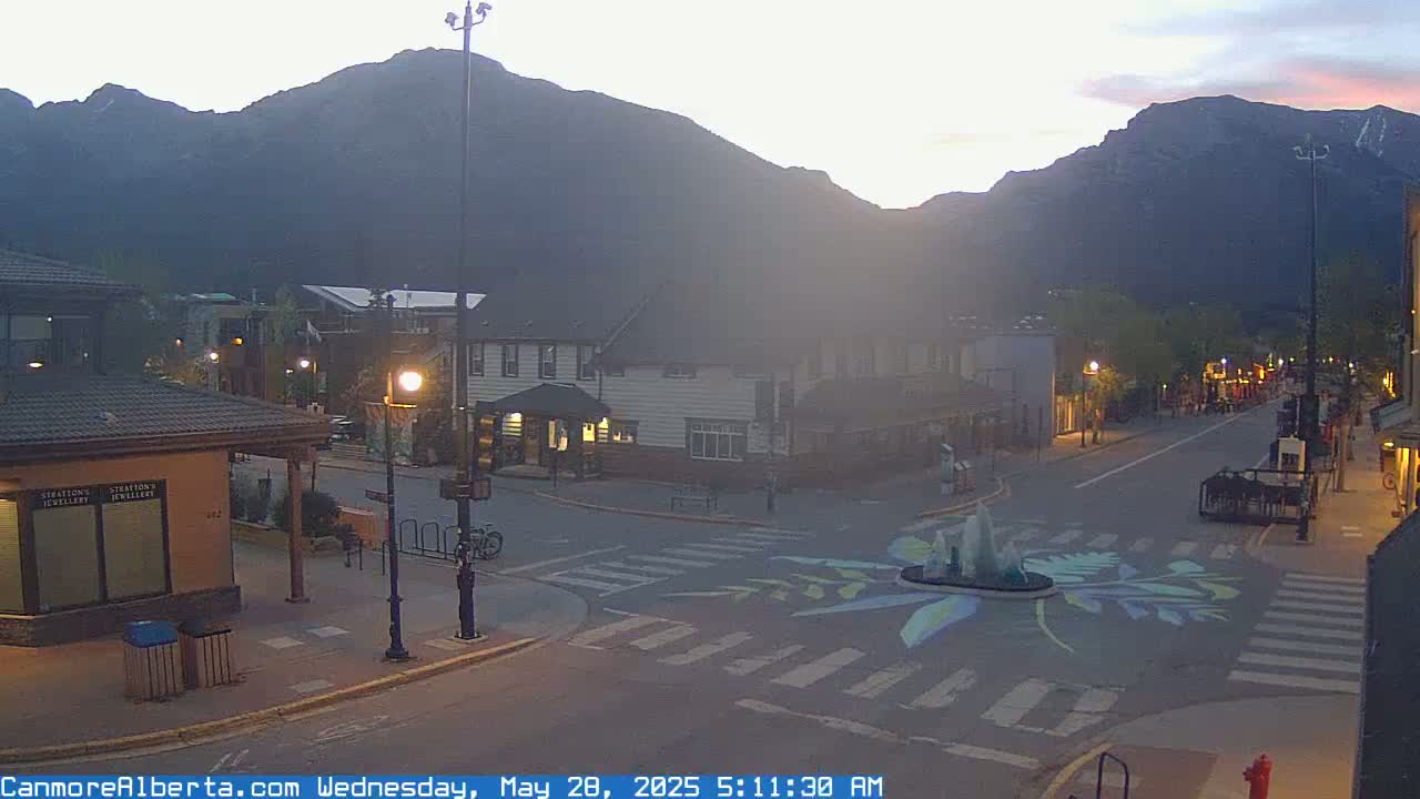 A mostly empty street in a mountain town at dawn, with a painted crosswalk design around a fountain.