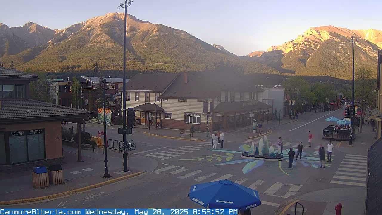 A street scene in a mountain town at sunset shows a street intersection with pedestrians and a small fountain, with buildings and mountains in the background under a clear sky.