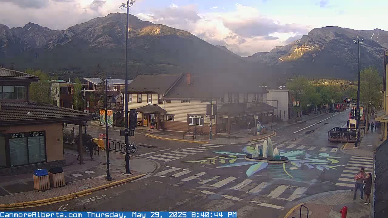 A street scene in a mountain town shows a crosswalk with a painted design in the center, buildings lining the street, and mountains in the background under partly cloudy skies.