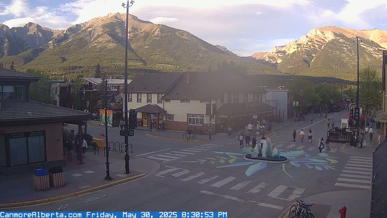 A town square with pedestrians and a fountain, situated in front of buildings under a partly cloudy evening sky, with mountains in the background.