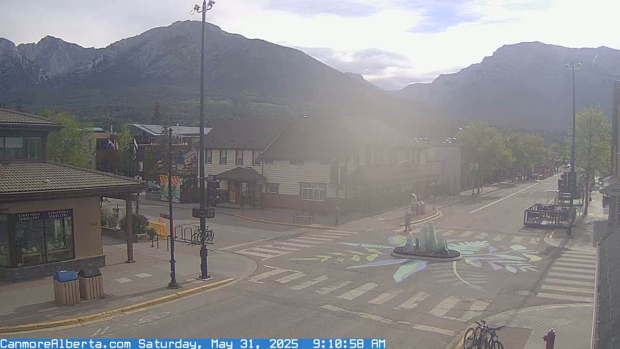 A hazy morning scene shows a mountain town street intersection with a painted crosswalk, buildings, and a few people and bicycles; mountains are visible in the background.